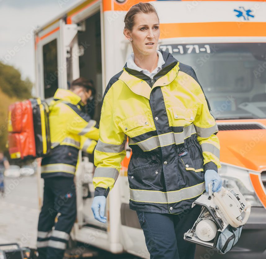 a paramedic carries equipment while another paramedic stands at an ambulance