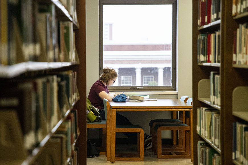 A student studies in the library with shelves full of books. 