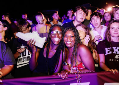 Two female students, one holding a Nike shoe, laugh at on the front row of the outdoor night time Flo Rida concert