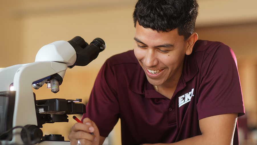 a smiling EKU student sits beside a microscope and makes notes