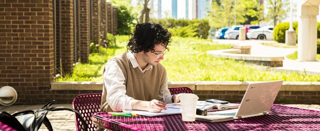 Male student sitting outside with laptop taking notes and studying