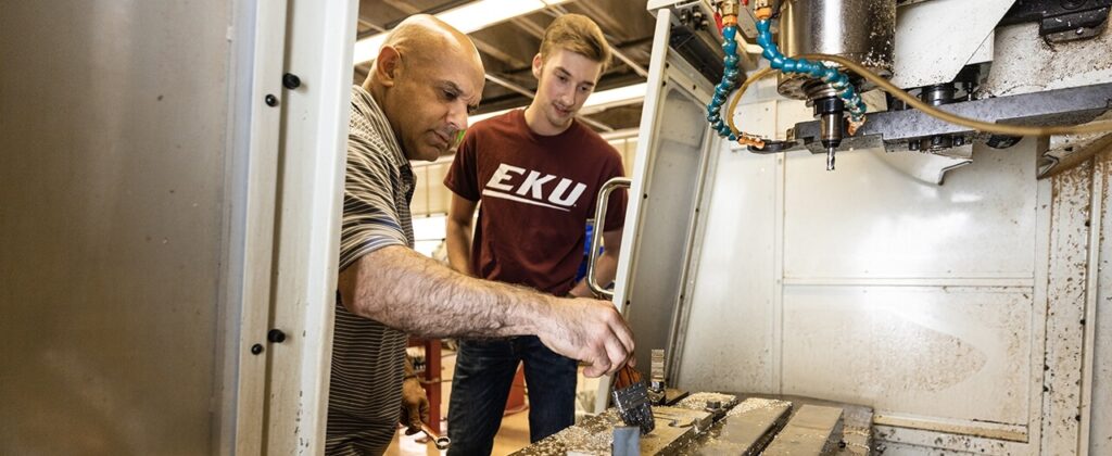 EKU manufacturing engineering student, in an EKU t-shirt, watches as a professor demonstrates cleaning of manufacturing equipment.