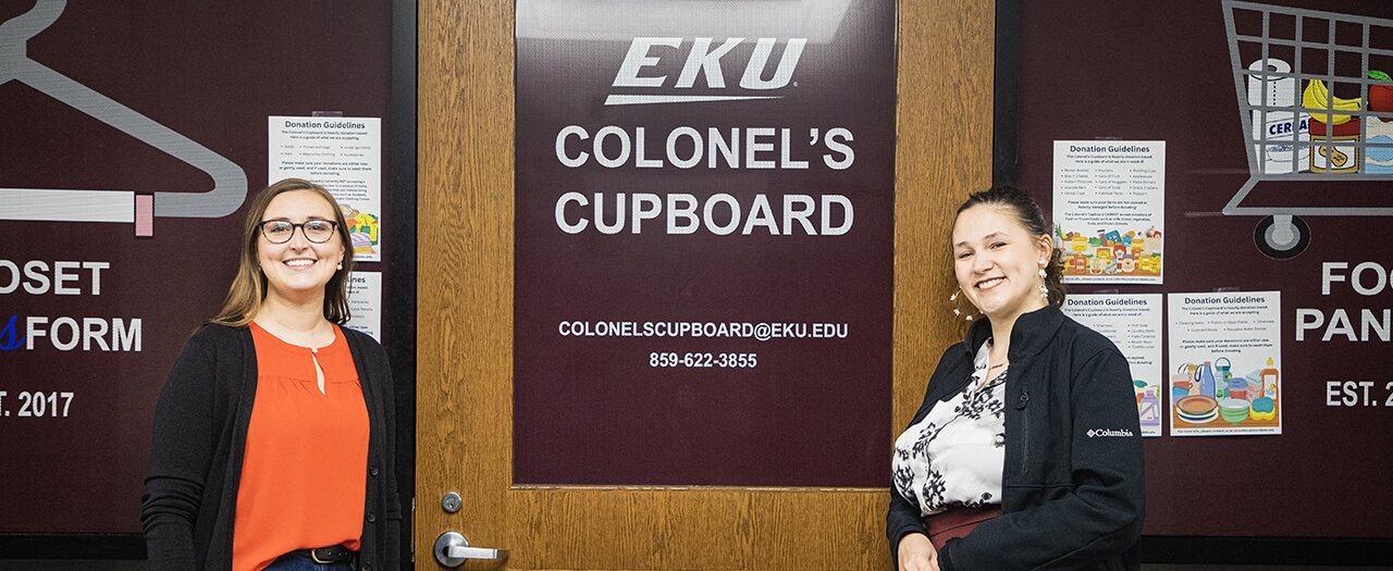 Two female EKU students pose in front of EKU's Colonel's Cupboard door.