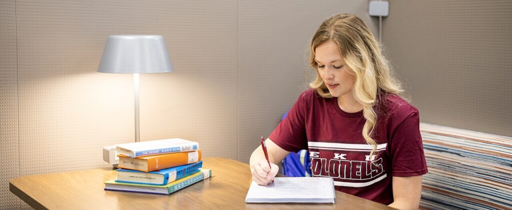 A female student wearing an EKU t-shirt sits at her desk with books and takes notes.