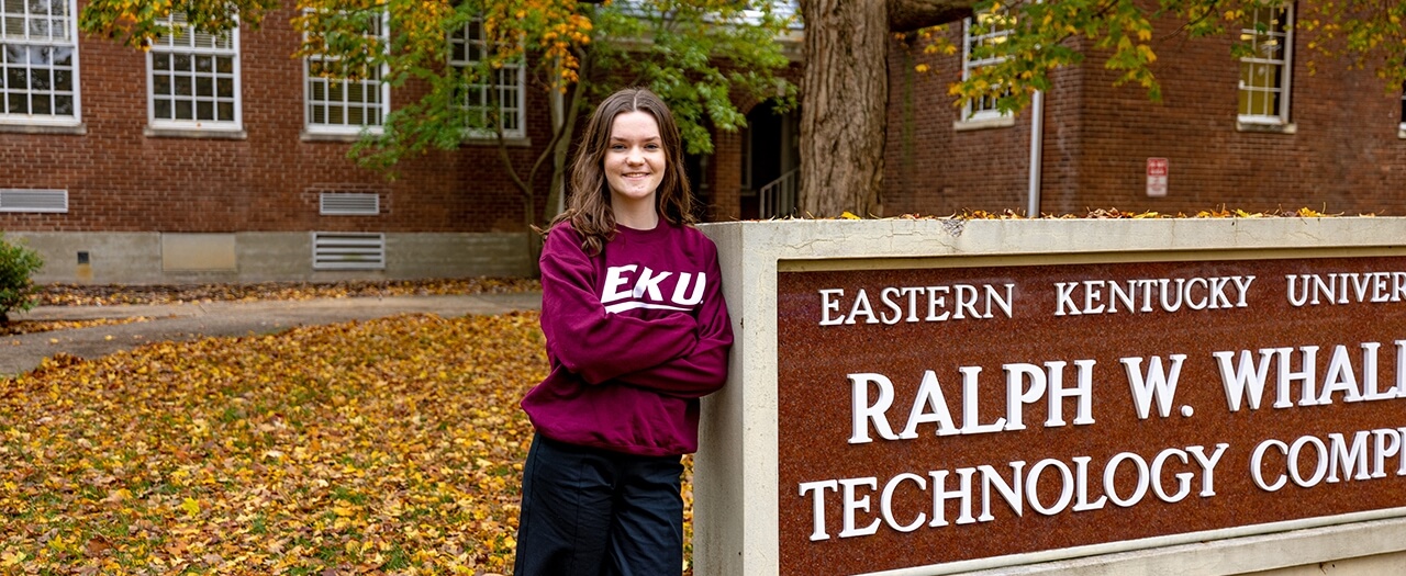 EKU manufacturing engineering student Jocelyn Langdorf, in an EKU sweatshirt, leans agains the Ralph W. Whalin Technology Complex sign on campus.