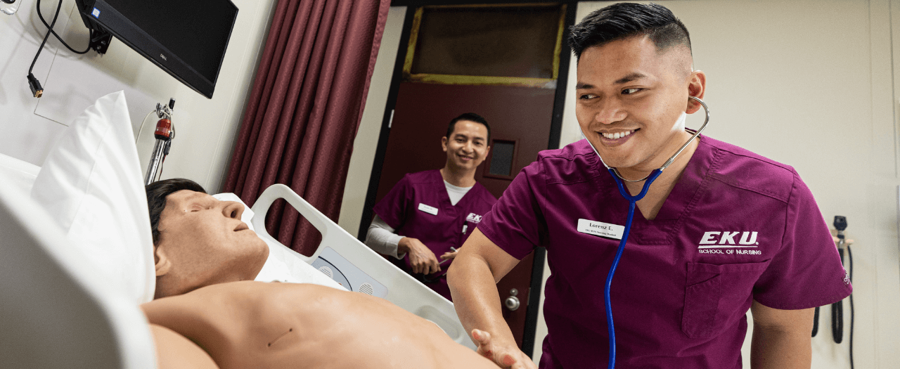 Two male EKU students in maroon scrubs check vitals on a training mannequin in a hospital setting.