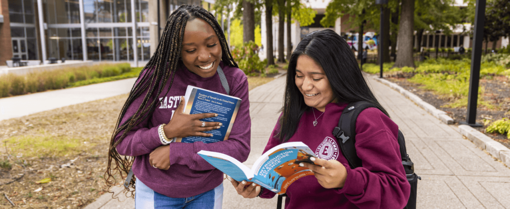 Two smiling female EKU students look into a book outside on campus.