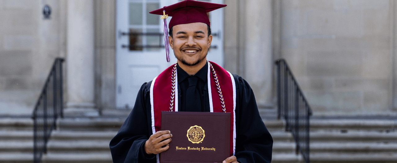 Bradley Lackey wearing his graduation regalia holds his diploma outside of Keen Johnson on EKU's campus.