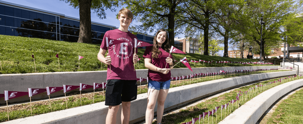 A male and female student hold EKU pennant flags amongst others staked in the ground to celebrate Giving Day 2023.