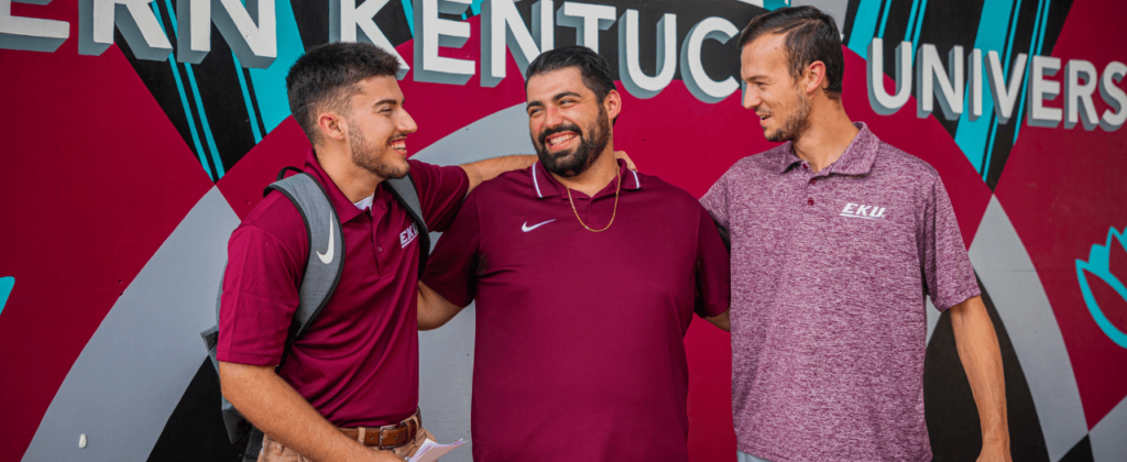 Three male EKU students share a laugh in front of the EKU mural outside of the Powell Building on campus.