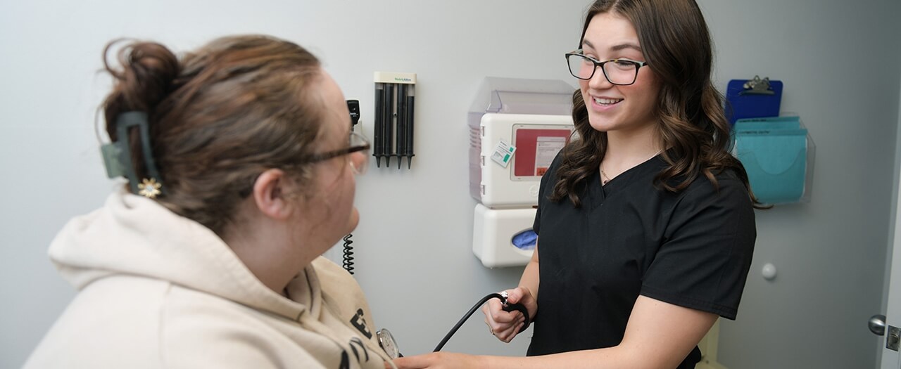 A female pre-med student in black scrubs checks a female patients blood pressure.