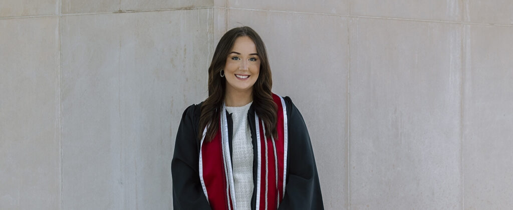 EKU graduate, Jenna Riel, in her graduation robe and cords, stands outside of a building on campus.