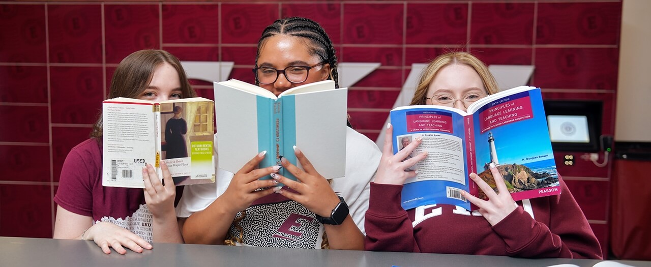 Three female students peek over their books while sitting at a table.