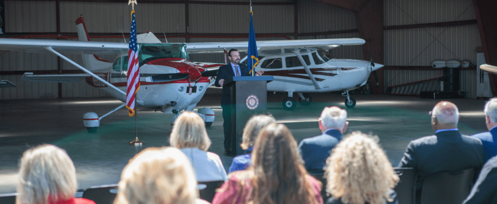 EKU President David McFaddin stands at a podium in front of a hanger with two small aircraft to address the crowd.