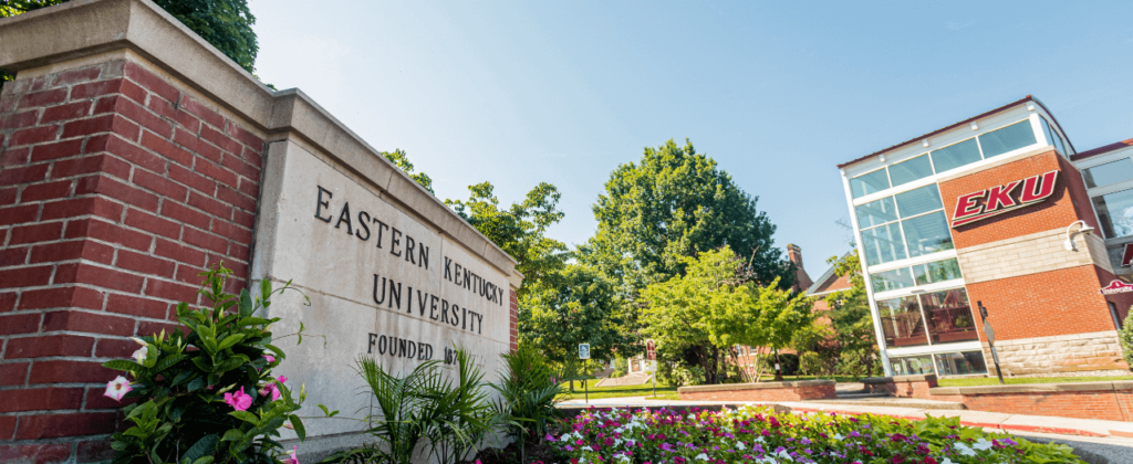 An upward view of the brick EKU sign and a spring flowerbed with the pedway tower in the background.