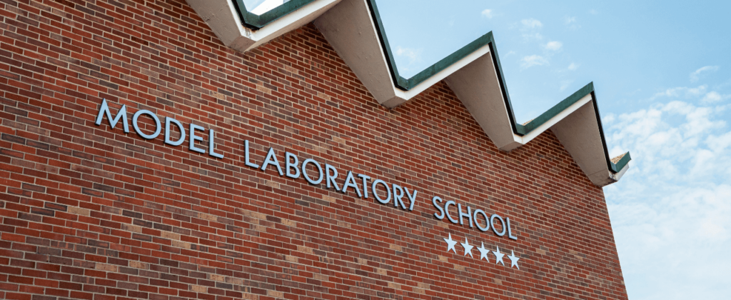 A close up of the red brick wall and corner of the roof of Model, with silver letters reading "Model Laboratory School" and five stars.