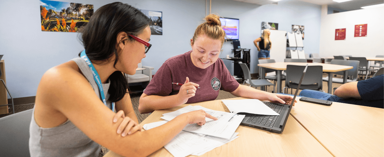 Two female EKU students work together at a table with notes and a laptop in the Student Success Center