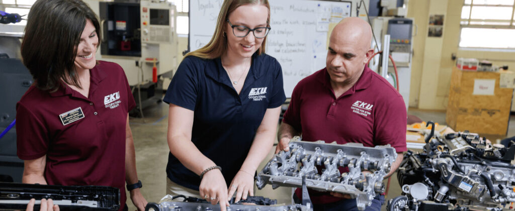 A female EKU student points out part of an auto engine to a male STEM faculty member and female Occupational Therapy faculty member in a auto shop setting.