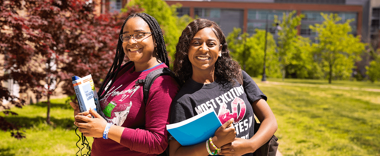 Two female EKU students hold their books outside on campus and stand back to back and smile.