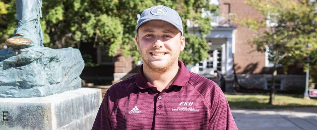 EKU Student Carson England wearing an EKU polo and standing next to EKU's Daniel Boone statue.