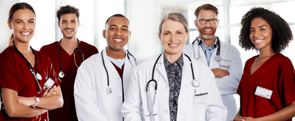 A group of medical professionals in scrubs and white coats smile while looking head on.