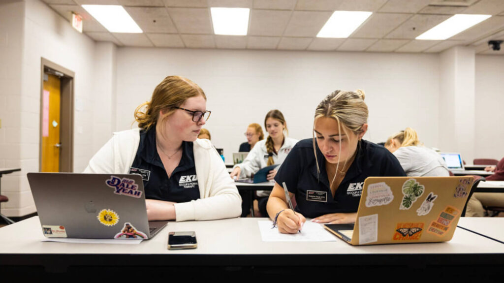 Two female OTD students in scrubs work together on an assignment at a desk in a classroom with their laptops open.