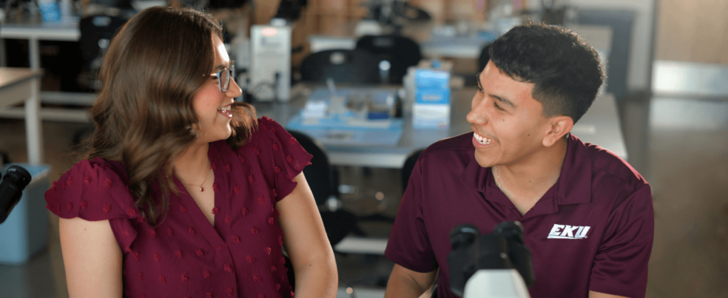 A female and male student, both in maroon, work with a microscope in a lab setting.