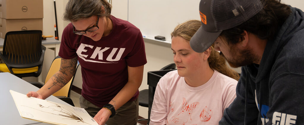 Dr. Sally Chambers, in an EKU t-shirt, shows materials to a female and male student at a desk.