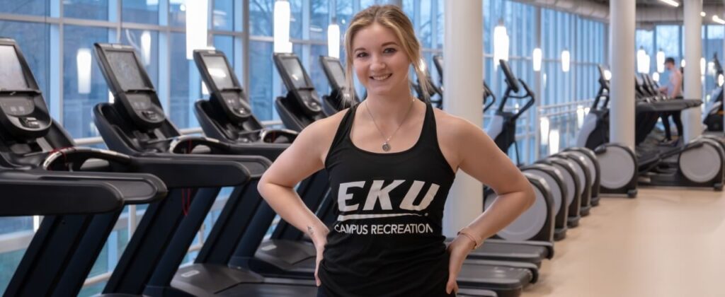 EKU student Julia Day, in a black EKU tank, standing in front of a row of treadmills at the Campus Recreation Center