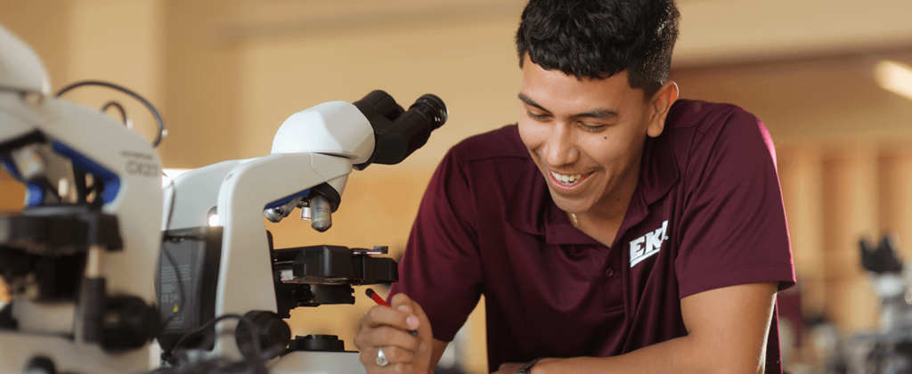 Dark haired student in EKU shirt takes notes near microscope in lab.