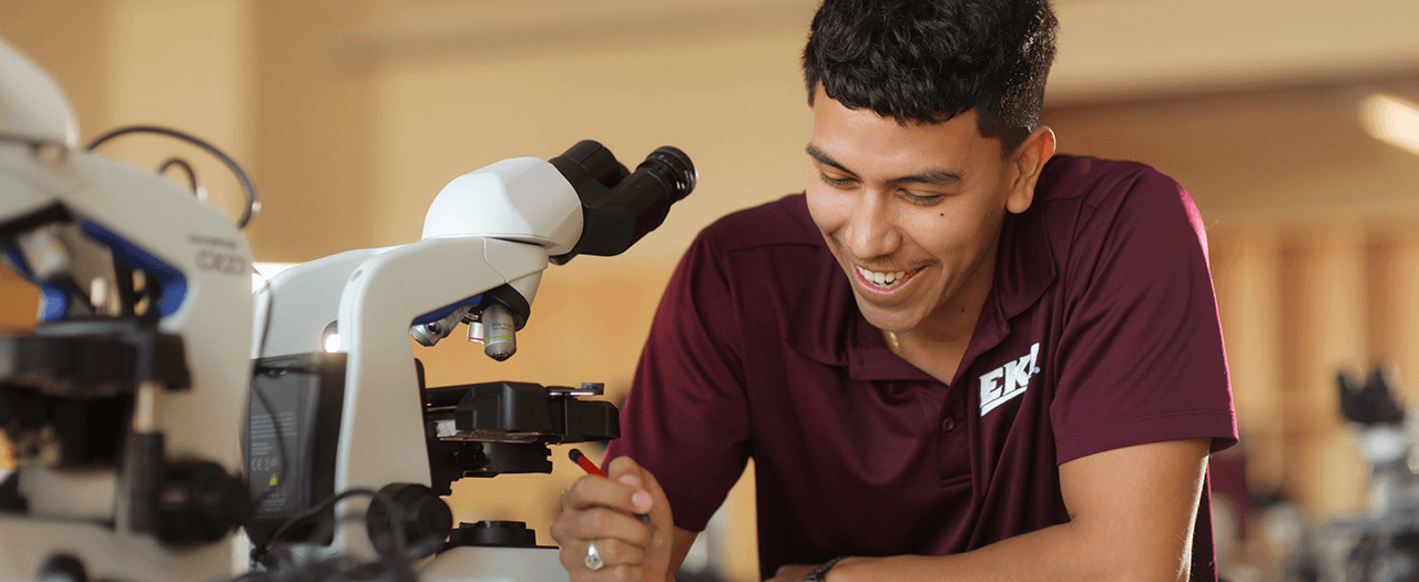 Dark haired student in EKU shirt takes notes near microscope in lab.