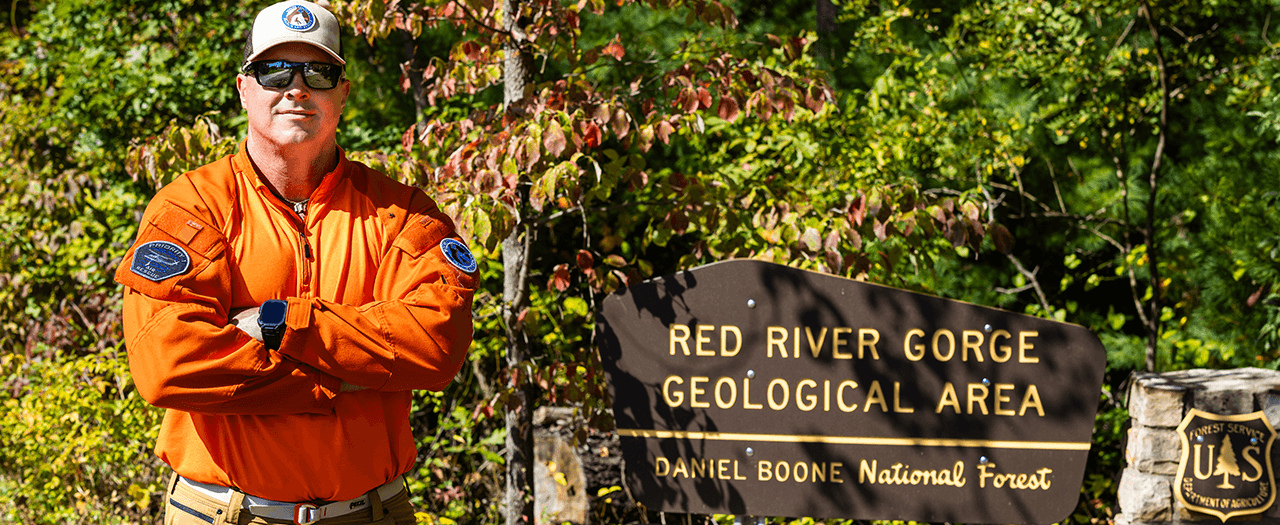 John May, wearing a bright orange uniform shirt, stands in front of the Red River Gorge sign in the Daniel Boone National Forest.