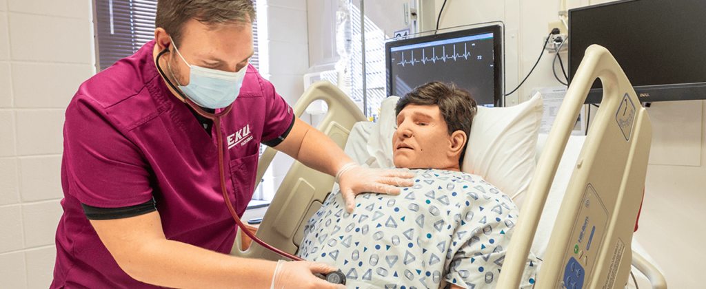 A male EKU nursing student in maroon scrubs practices with a mannequin in a hospital setting on campus.