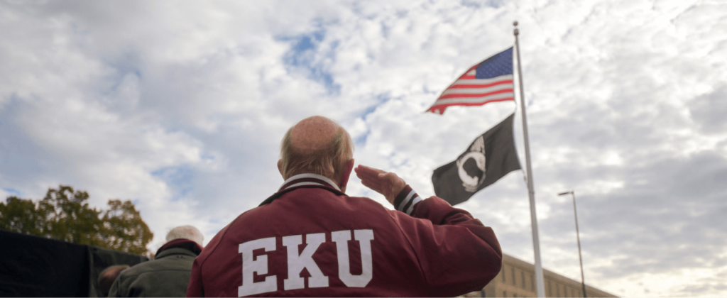 EKU veteran in a vintage satin EKU jacket salutes the American flag at the Veterans Memorial.