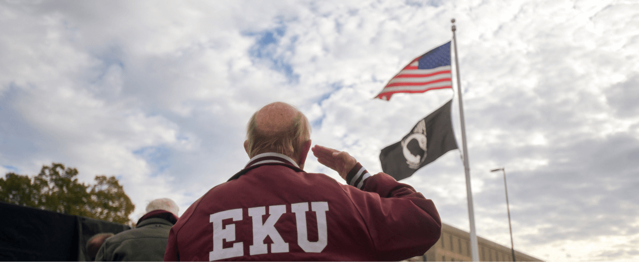 EKU veteran in a vintage satin EKU jacket salutes the American flag at the Veterans Memorial.