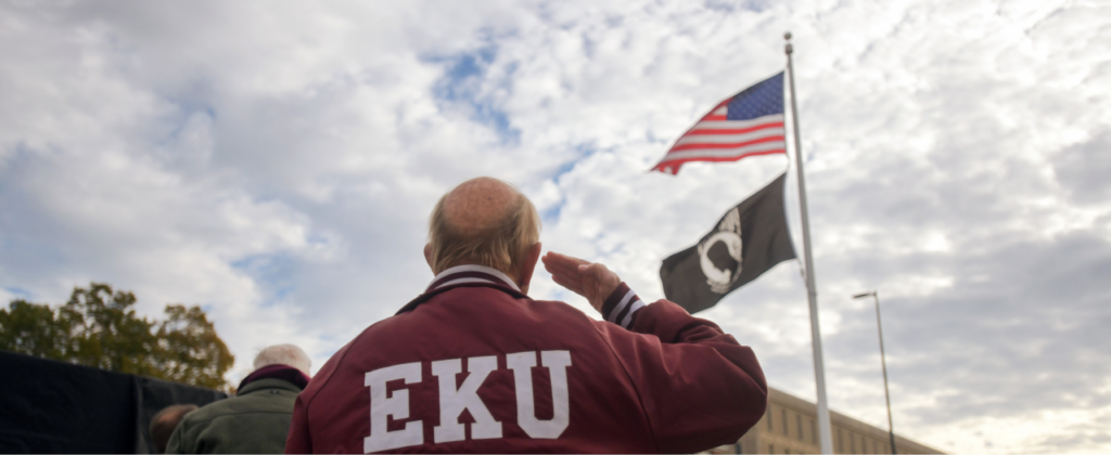 a veteran wearing an EKU jacket salutes the American and POW flags