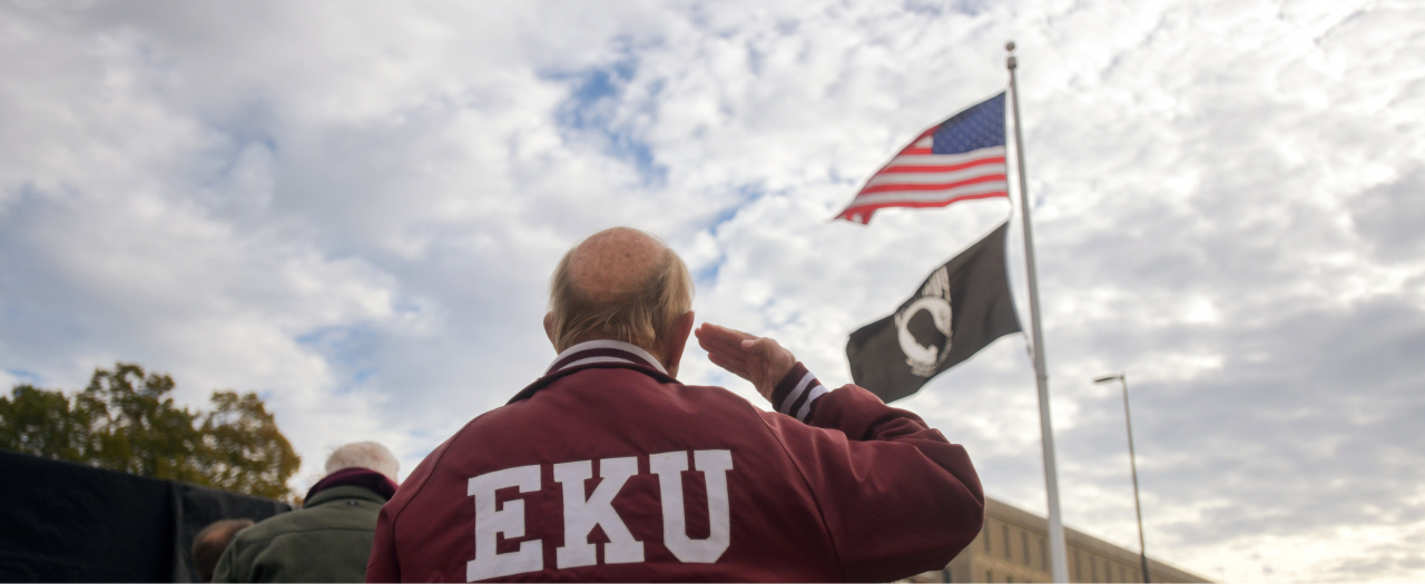 a veteran wearing an EKU jacket salutes the American and POW flags