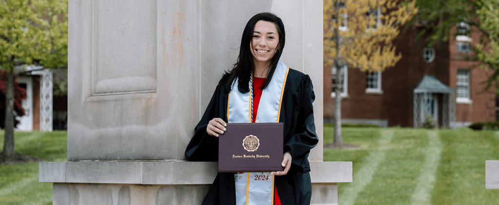 EKU graduate, Emma Thomas, holds her degree while wearing her graduation regalia outside on EKU's campus.