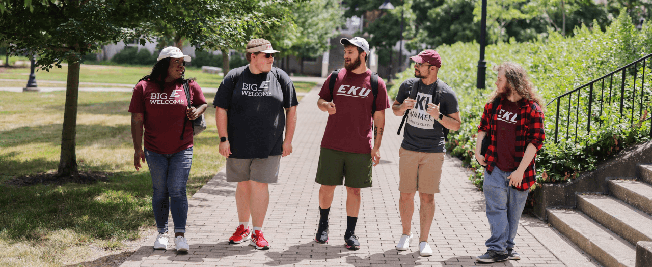 A line of five students, wearing EKU gear, walking on a sidewalk on EKU's campus and talking.