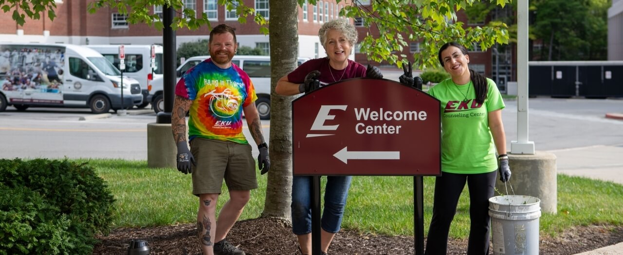 Three EKU employees, wearing EKU shirts and gloves, take a break from mulching near the welcome center to smile.