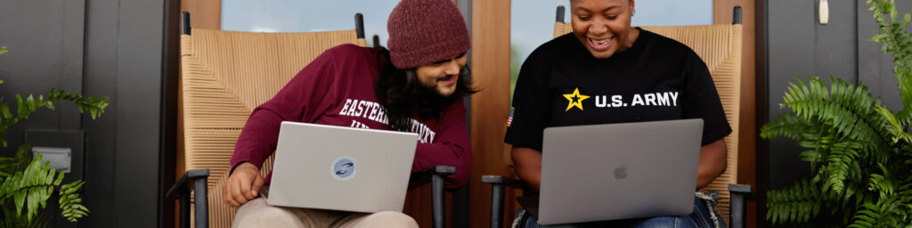 Two EKU students, one male and one female, sit on a porch with their laptops discussing an assignment.