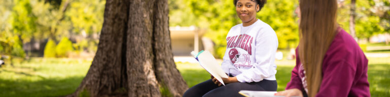 Two female students sit in the ravine on EKU's campus, with open books, during a study session.