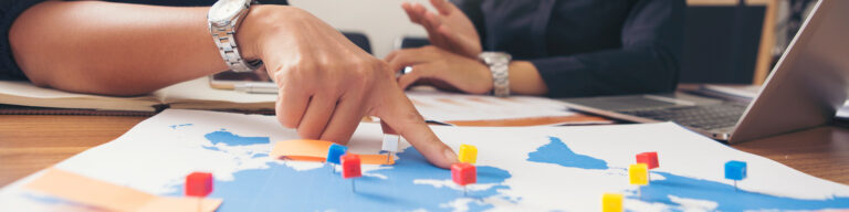 a hand points to different colored pins on a map of the world with another pair of hands on a chart in the background