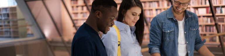 three students smile as they stand together looking down at a table in a library