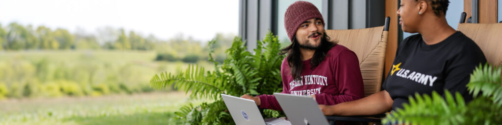 A male student in an EKU shirt and a female student in a U.S. Army shirt sit on the front porch with their laptops open, discussing coursework.