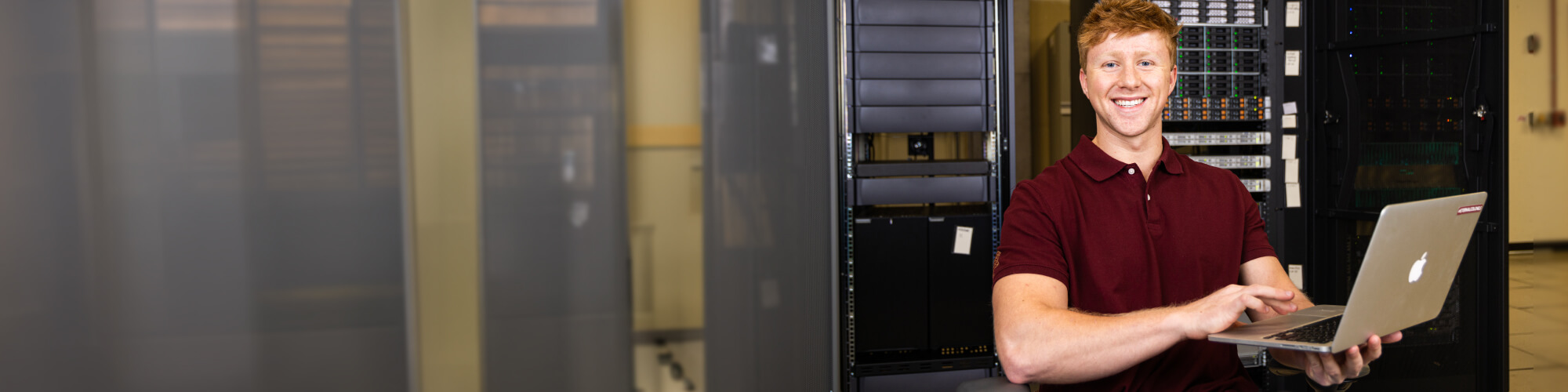 a smiling student uses a laptop while sitting in front of a bank of network servers