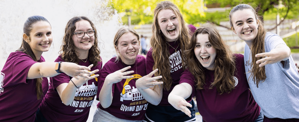 Six female EKU students, all wearing different EKU t-shirts, hold up the "E" hand signal in front of the fountain in Powell Plaza.