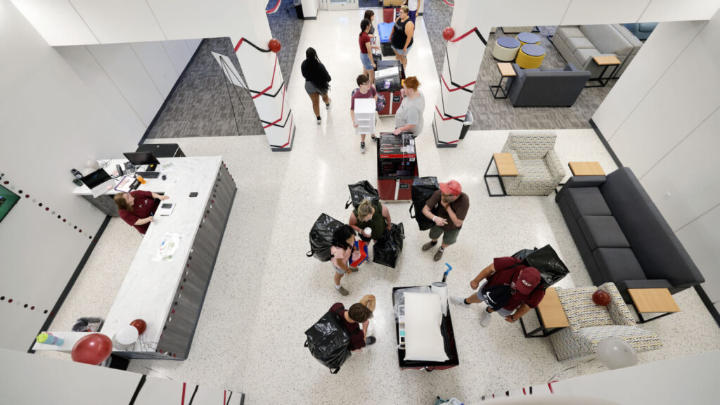 Students bring their belongings into the lobby of Keene Hall on move-in day. The front desk is in view along with couches and chairs and tables.