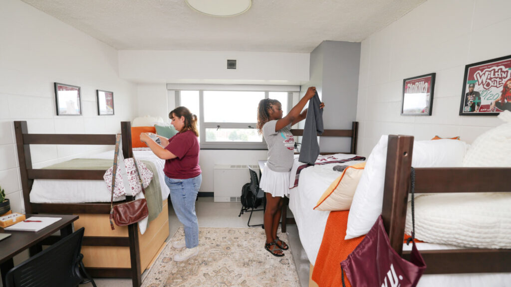 Two students unpack on the lofted beds in a shared room in Keene Hall. Windows and one of two desks are in view.