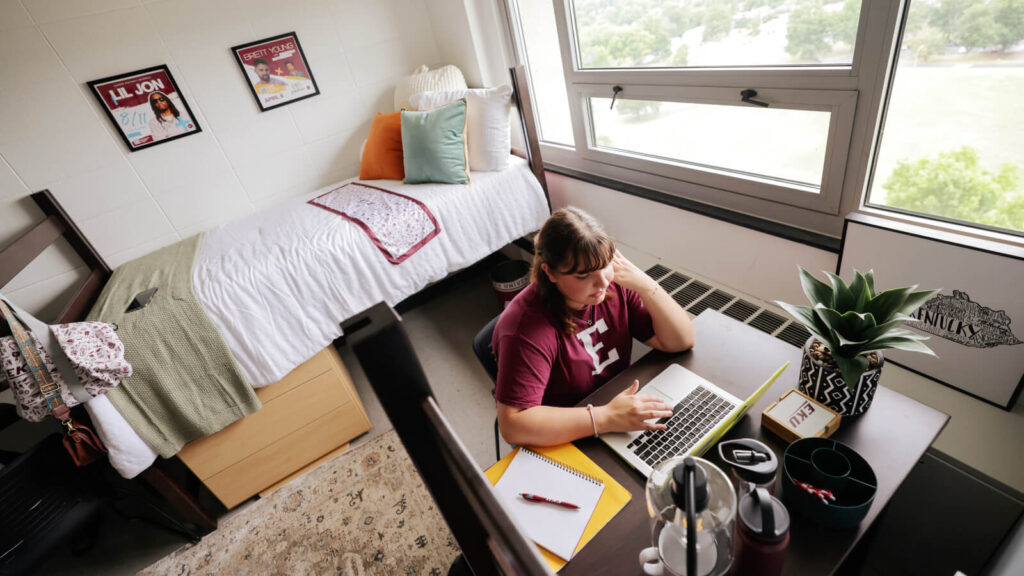 A student sits at a study table using a laptop in a shared room in Keene Hall. A lofted bed beside windows is in view.